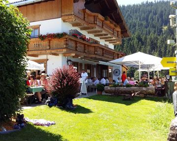 Ein gemütliches Restaurant mit Sonnenschirmen und Gästen im Freien, umgeben von grünen Wiesen und Bergen. Die Fassade des Hauses ist im traditionellen Stil mit Blumenkästen dekoriert.