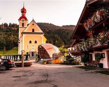 Eine malerische Dorfstraße mit einer Kirche und traditionellen Holzgebäuden. Im Hintergrund sind bewaldete Hügel zu sehen.