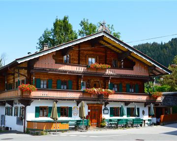 A traditional wooden house in alpine style with green shutters and blooming balconies. It is located surrounded by trees and a clear blue sky.