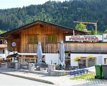 Eine gemütliche Pizzeria mit Holzfassade und Außenterrasse. Umgeben von grünen Bergen und einer ruhigen Straße.