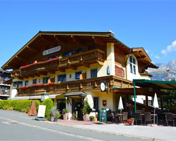 Ein charmantes alpines Hotel mit Holzverzierung und Balkonen. Im Hintergrund sind schöne Berge und ein klarer blauer Himmel zu sehen.