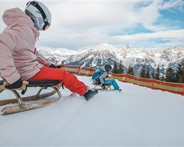 Zwei Kinder sitzen auf Schlitten im Schnee. Im Hintergrund sind schneebedeckte Berge und ein blauer Himmel zu sehen.