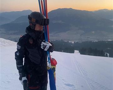 A skier stands on a snowy slope with ski poles in hand. In the background, the mountains and the sunset can be seen.