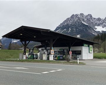 Eine Tankstelle mit einem überdachten Bereich in einer ländlichen Umgebung. Im Hintergrund sind beeindruckende Berge zu sehen.