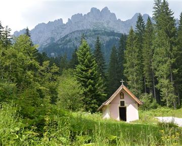 Eine kleine Kapelle steht am Rande eines Waldes, umgeben von hohen Bäumen. Im Hintergrund erheben sich majestätische Berge.