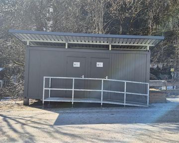 A modern, gray changing room stands in a paved area. Surrounded by trees and a clear sky, it appears inviting and functional.