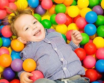 A smiling child is lying in a colorful ball pit. The colorful balls are in different colors such as yellow, blue, and red.