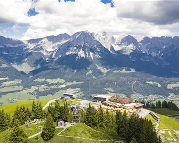An impressive mountain landscape with high peaks and green meadows. In the foreground, there are trees and buildings that complement the natural beauty of the surroundings.