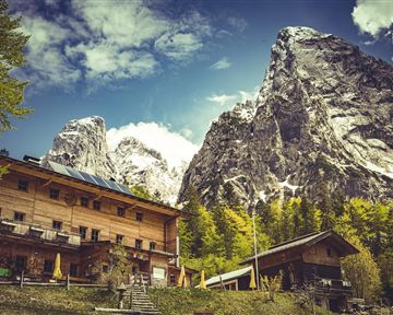 Eine malerische Berglandschaft mit hohen Gipfeln und einer charmanten Holzunterkunft im Vordergrund. Der Himmel ist teilweise bewölkt und die Natur ist üppig grün.