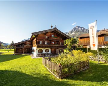 A traditional alpine wooden house with a lovingly designed garden. In the background, majestic mountains and a clear blue sky can be seen.