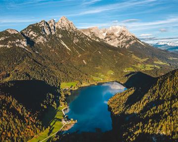 A breathtaking mountain landscape with snow-covered peaks and a clear lake in the foreground. Nature is surrounded by green forests and fields.