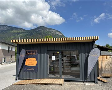 Ein modernes Geschäft aus Containern in einer ländlichen Umgebung. Im Hintergrund sind Berge und ein blauer Himmel zu sehen.