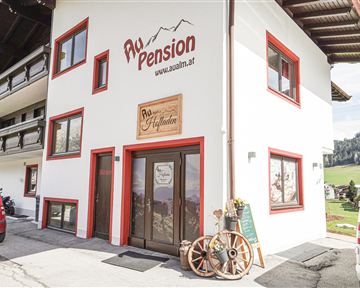 A cozy guesthouse with red window trim and an inviting entrance. In the background, green hills and forests can be seen.