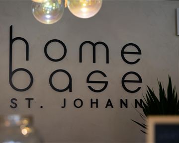 Ein modernes Schild mit der Aufschrift "home base St. Johann" an einer Wand. Im Hintergrund ist eine Pflanze und eine warme Lichtquelle zu sehen.