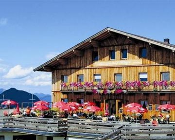 A rustic wooden house with balconies adorned with flowers. In the foreground, there are tables with red umbrellas, surrounded by a picturesque mountain landscape.