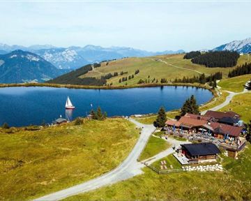 A picturesque landscape with a clear lake and a cozy mountain house. In the background, green meadows stretch out and the mountains of the Alps rise.