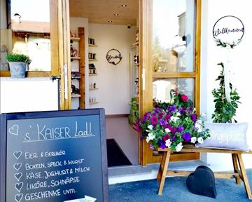 Ein einladender Geschäftsausgang mit Holztüren und bunten Blumen. Im Vordergrund steht ein Schild mit dem Angebot von frischen Produkten und Geschenken.