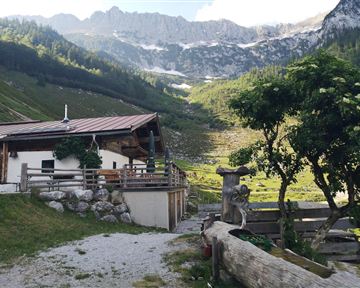 Eine gemütliche Hütte inmitten einer malerischen Berglandschaft. Im Hintergrund sind grüne Wiesen und hohe Berge zu sehen.