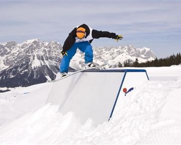 Ein Snowboarder springt über ein Hindernis in einer verschneiten Berglandschaft. Im Hintergrund sind mächtige Berge und ein blauer Himmel zu sehen.