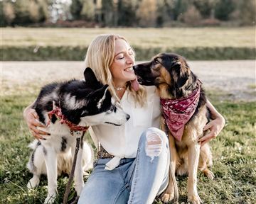 Eine Frau sitzt im Gras und umarmt zwei Hunde. Sie lächelt glücklich, während die Hunde liebevoll bei ihr sind.
