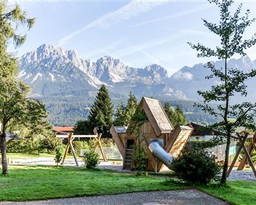 A playground landscape with a creative wooden play structure and swings, surrounded by green meadows. In the background, majestic mountains rise under a clear sky.