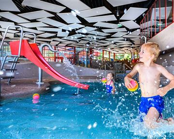 A children's pool with a red slide. Two kids are playing in the water and splashing around happily.