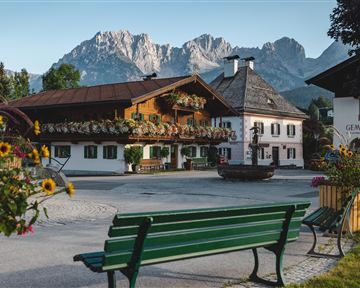 A picturesque village square with traditional wooden houses and blooming flowers. In the background, majestic mountains rise under a clear sky.