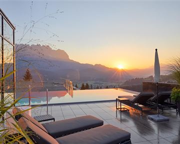 A beautiful pool area with loungers and a view of the mountains. The sun sets behind the mountains, bathing the scene in warm light.