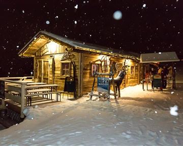 Eine gemütliche Berghütte im Schnee bei Nacht, mit festlicher Beleuchtung. Im Vordergrund sind Tische und eine Figur eines Rehs sichtbar.