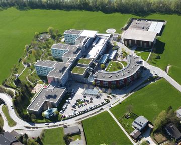 Ein modernes Gebäudeensemble in einer grünen Landschaft. Die Struktur umfasst mehrere separate Gebäude und ist von Rasenflächen umgeben.