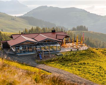 A modern building in the mountains with a terrace and wide green spaces. In the background, gentle hills and a clear sky can be seen.