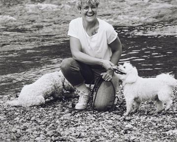 Eine fröhliche Frau kniet am Ufer eines Gewässers und hält zwei kleine Hunde an der Leine. Im Hintergrund sind Steine und Wasser sichtbar.
