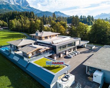 A modern building in a rural setting, surrounded by trees and mountains. There are parking spaces and a large meadow in the foreground.