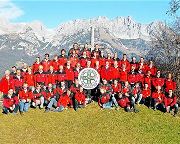 Eine große Gruppe von Menschen in roten Jacken steht auf einer Wiese vor majestätischen Bergen. Sie posieren um ein gemeinsames Schild in der Mitte des Bildes.