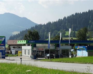 Eine OMV-Tankstelle umgeben von Bergen und Bäumen. Der Himmel ist klar und die Landschaft ist grün.