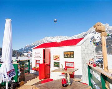 Ein gemütliches Holzhaus mit rotem Dach in einer schneebedeckten Berglandschaft. Der Himmel ist klar blau und verschiedene Leuchten schmücken den Bereich.