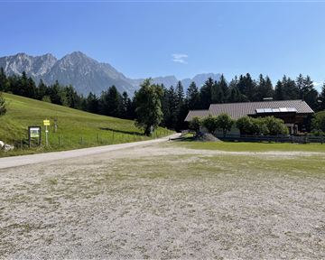 Eine idyllische Landschaft mit sanften Hügeln und hohen Bergen im Hintergrund. Im Vordergrund steht ein typisches alpenländisches Gebäude.
