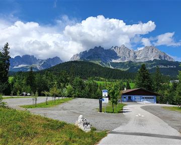 Ein malerischer Parkplatz mit Blick auf majestätische Berge und eine grüne Landschaft. Der Himmel ist teilweise bewölkt und blau.