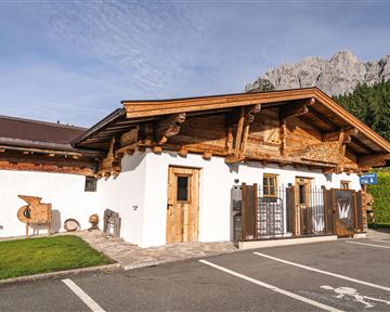 A traditional chalet with wooden cladding in a picturesque setting. In the background, you can see impressive mountains and a green meadow.