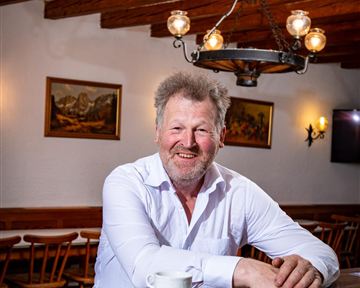 Ein Mann mit einem freundlichen Lächeln sitzt an einer Holztheke. Auf der Theke steht eine Tasse, und im Hintergrund sind gemütliche Sitzplätze zu sehen.