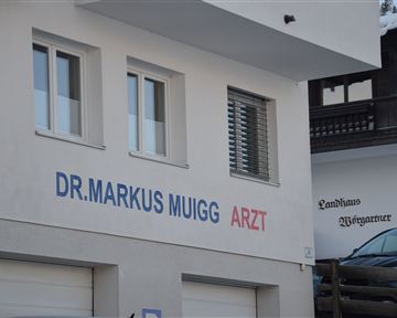 Das Bild zeigt die Außenansicht einer Arztpraxis mit dem Schriftzug "Dr. Markus Muigg Arzt". Die Fassade ist schlicht und modern gestaltet.