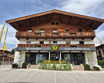 A modern bank branch in alpine style with wooden cladding. In front of the bank, there are yellow flags and there is green planting.