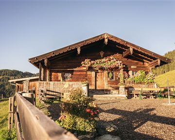Eine rustikale Hütte aus Holz umgeben von grünen Wiesen und Bergen. Der Himmel ist klar und die Landschaft wirkt friedlich.