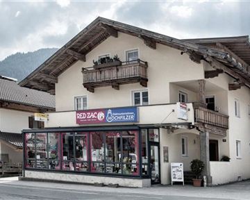 Ein traditionelles Gebäude mit einem Geschäft im Erdgeschoss und Balkonen im Obergeschoss. Die Umgebung zeigt ländliche Architektur und eine bergige Kulisse.