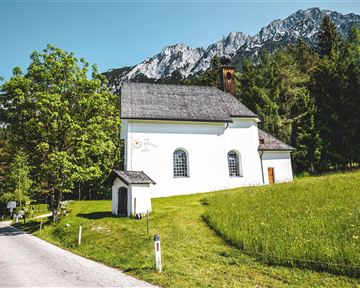 A small, white church stands idyllically in a green meadow. In the background, majestic mountains can be seen, and the sky is clear.