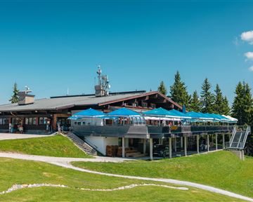 A modern restaurant in the mountains with a large terrace and blue sun umbrellas. Surrounded by green meadows and coniferous trees under a clear blue sky.