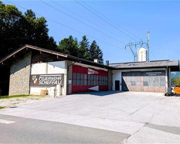 A fire station in Scheffau with a red garage door. The surroundings are surrounded by trees and the sky is clear and blue.