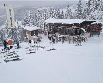 Eine verschneite Landschaft mit einem Skihütten und vielen Skiern, die abgestellt sind. Im Hintergrund sind hohe, schneebedeckte Bäume zu sehen.