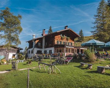 Ein rustikales Berghaus umgeben von einer grünen Wiese und Bäumen. Im Vordergrund sind Sitzmöglichkeiten und Sonnenschirme zu sehen.