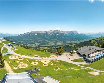 Eine malerische Berglandschaft mit grünen Wiesen und schneebedeckten Gipfeln im Hintergrund. Im Vordergrund sind Spielplätze und Gebäude zu sehen, die eine schöne Aussicht bieten.